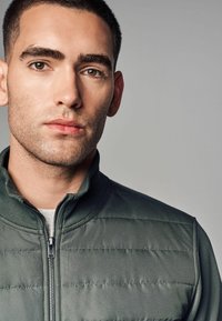 Grün gesteppte Jacke mit hohem Kragen und einem Reißverschluss auf der Vorderseite. Das Material wirkt glatt und weist horizontale Nähte über die Brust auf.