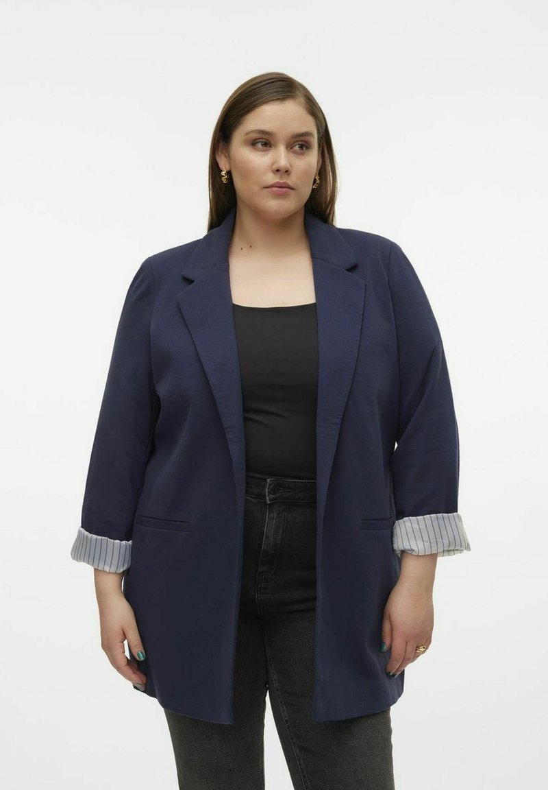 Navyblazer mit strukturierten Schultern, offenem Frontalverschluss, zwei Fronttaschen und gestreiften Akzenten an den Bündchen. Über einem schwarzen Oberteil und dunklen Jeans getragen.