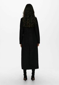 Long black coat in a smooth fabric, straight design with long sleeves, features a side split, and paired with high-heeled black boots.