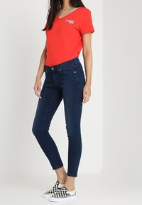 Camiseta roja de cuello en V con el logo "TOMMY", pantalones vaqueros ajustados de mezclilla oscura y zapatos de lona a rayas en blanco y negro. Texturas de algodón y mezclilla suaves.