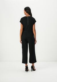 Svart jumpsuit med korte ermer, glidelås bak, vide cropped ben og en teksturert stoff. Brukt med svarte høyhælte sko.