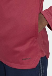 Camiseta deportiva roja AEROREADY con tejido texturizado, mangas cortas y bolsillos laterales, combinada con pantalones azul oscuro con rayas.
