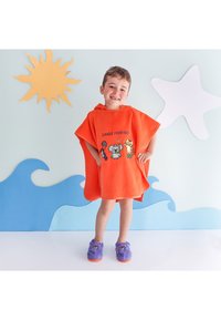 Smiling child in orange jungle-themed hooded towel and purple slippers stands against wall decorated with sun, starfish, and waves.