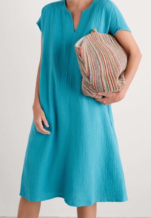 Osoba ubrana w turkusową sukienkę o fakturze, sięgającą do kolan, trzymająca dużą, wielokolorową, paskowaną plecioną kopertówkę na jednolitym tle.