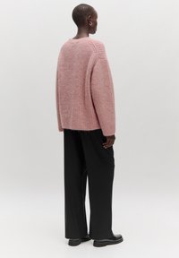 Rosa gestrickter Pullover mit lockerer Passform, gerippter Textur und tiefem Schulterbereich, kombiniert mit schwarzen weit geschnittenen Hosen und glänzenden schwarzen Schuhen.