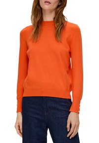 Oranges Strickpullover mit rundem Halsausschnitt und langen Ärmeln, die an den Bündchen mit Knopfakzenten versehen sind, kombiniert mit dunkelblauen Jeans.