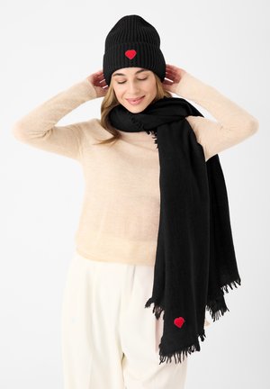 Schwarze, gerippte Mütze mit einem roten Herz-Emblem, kombiniert mit einem großen schwarzen Schal, getragen über einem hellbeigen Pullover. Glatte Texturen, lässige Passform.