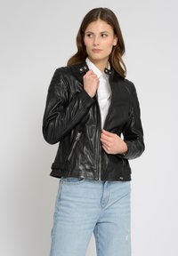 Schwarze Lederjacke mit tailliertem Design, Reißverschluss vorne, Druckknopfkragen und Seitentaschen. Getragen über einem weißen Hemd und hellblauen Jeans.