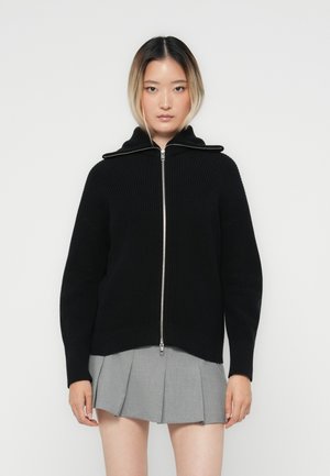 Maglione nero lavorato a maglia con zip e colletto alto, trama a coste e hardware in argento. Il modello indossa una gonna a pieghe grigia.