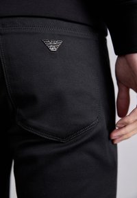 Black denim pants with a textured finish, featuring a silver logo on the back pocket and fine stitching along the seams.
