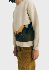 Maglione di lana beige con una sezione inferiore a motivo verde e giallo, polsini a coste e una toppa con logo sull'orlo. Abbinato a pantaloni in corduroy marroni.
