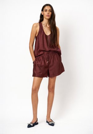 Vrouw staand, draagt een mouwloze kastanjebruine top en bijpassende shorts met zwarte puntige slingback schoenen met strikaccenten.