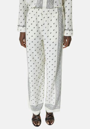 Białe spodnie z czarnym wzorem paisley i wzorem na obrzeżach, noszone z pasującym topem i czarnymi sandałami na ramiączkach na osobie stojącej.