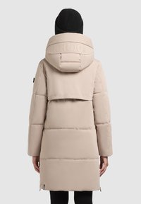 Manteau matelassé beige clair avec capuche, présentant un design matelassé, un logo à l'arrière et des fermetures éclair sur le côté. Bonnet en tricot texturé visible.