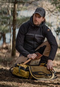 Uomo con giacca nera e cappellino si inginocchia all'aperto, tenendo un'ascia e aprendo uno zaino da campeggio giallo sul terreno della foresta.