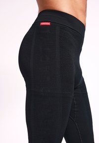 Leggings deportivos negros con un diseño de punto texturizado, cintura alta y detalle de logo en una etiqueta roja prominente. Forma ajustada.