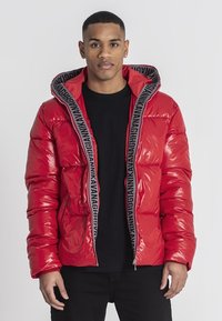 Rote Pufferjacke mit glänzender Oberfläche, schwarzem Logo-Detail am Reißverschluss und Kapuze. Hat eine kastige Form und ein gesteppte Design.