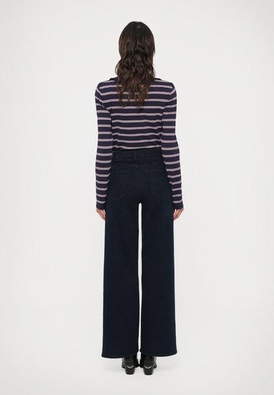 Triibuline pikkade varrukatega topp ümara kaelusega, tumesinised laiade säärega teksapüksid ja mustad kõrge kontsaga kingad, vaadatuna tagant valgel taustal.