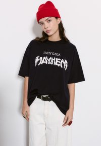 Musta ylipitkä t-paita, jossa valkoinen graafinen teksti "LVDY GAGA MAYHEM". Mallilla on päällään kermaiset leveälahkeiset housut ja punainen ribbikuvioinen pipo.