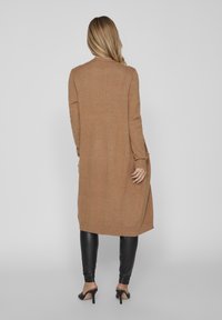 Cardigan lungo di colore beige realizzato in un materiale morbido, con polsini e orlo a costine, abbinato a leggings neri eleganti e tacchi alti.