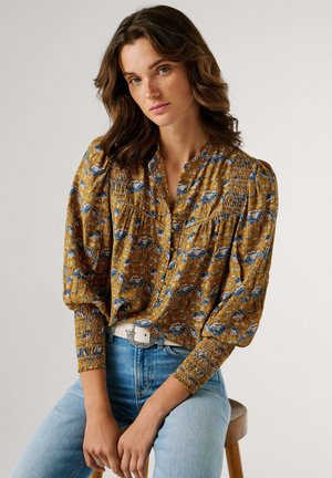 Blusa de color mostaza con estampado floral azul, mangas abullonadas y puños fruncidos. Combinada con jeans azul claro y un cinturón blanco.