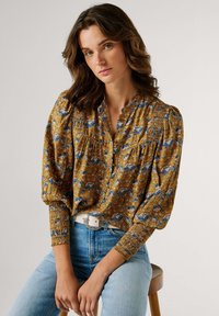 Senfgelbe Bluse mit blauem Blumenmuster, Puffärmeln und elastischen Bündchen. Kombiniert mit hellblauen Jeans und einem weißen Gürtel.