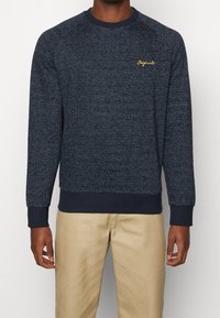 Marineblauer Sweatshirt mit strukturiertem Muster, geripptem Kragen und Bündchen sowie kleinem goldenen "Originals"-Logo auf der Brust. Getragen mit beigen Hosen.