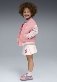Chaqueta varsity rosa con mangas blancas, logo bordado, combinada con una falda rosa claro con un gráfico colorido. Zapatillas rosas con acentos.