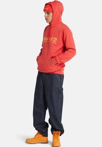 Sudadera roja con capucha y logo amarillo, pantalones de mezclilla azul sueltos y botas naranjas. Presenta un bolsillo tipo canguro y puños acanalados en la sudadera.