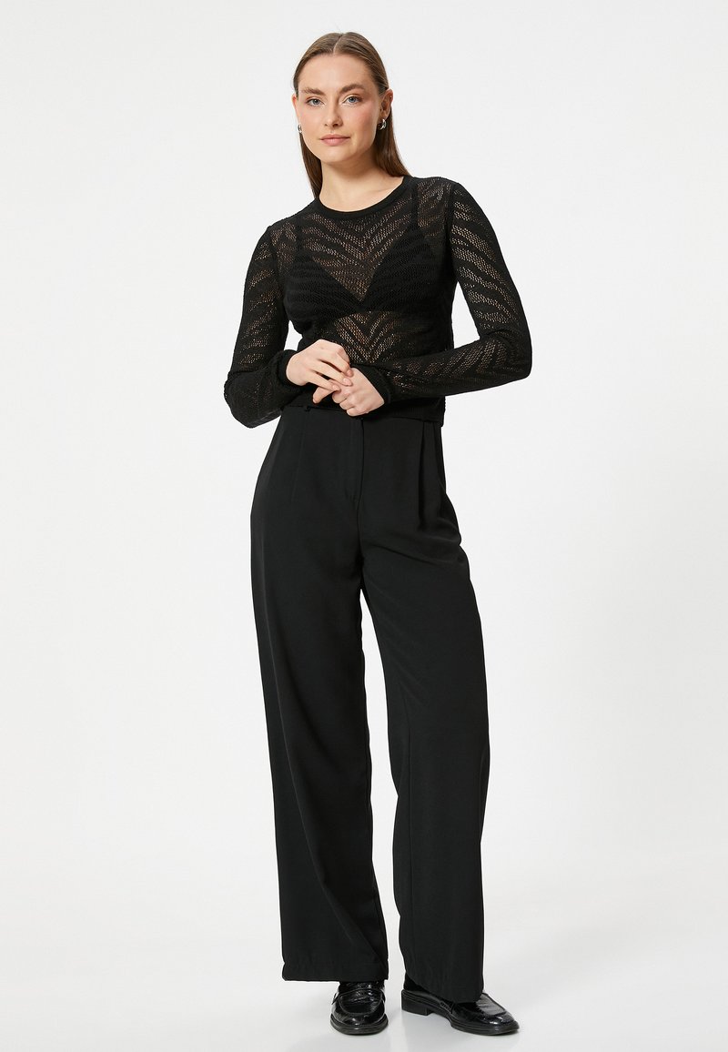 Top in rete nera a maniche lunghe con motivo a chevron, abbinato a pantaloni neri a vita alta e a gamba larga e scarpe nere, in posa contro uno sfondo neutro.