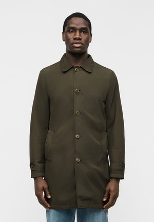 Mand iført en kneppet, mørkeolivengrøn frakke med forlommer og blå jeans, stående foran en ensfarvet lysegrå baggrund.