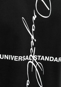 Czarna tkanina z białym napisem „UNIVERSAL STANDARD”, częściowo zakryta pionowym białym kursywnym pismem przebiegającym przez nią.