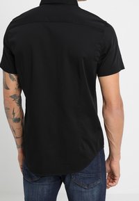 Camisa negra de manga corta con cuello estructurado, tela suave, dobladillo curvado y diseño liso en la parte trasera. Se lleva con pantalones vaqueros azules.