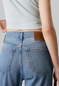 Jasnoniebieskie dżinsy o wysokim stanie z brązową skórzaną metką Calvin Klein z tyłu. Elementy to wysokie biodra oraz dwie tylne kieszenie ze stębnowaniami.