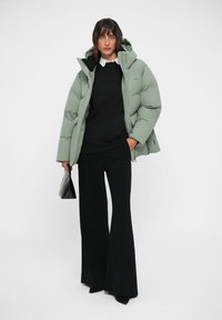 Puffer jacket in light green, featuring a hood and quilted design. Worn over a black sweater with a white collar, paired with black flared pants.