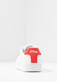 Sneaker bianco con accenti rossi, tomaia texturizzata e collo imbottito. Logo visibile sul tallone. Suola bianca spessa con una finitura leggermente texturizzata.
