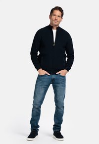 Dunkelblauer Reißverschluss-Pullover mit gerippter Textur, hohem Kragen und goldenem Reißverschluss. Kombiniert mit hellblauen Jeans und schwarzen Sneakers.