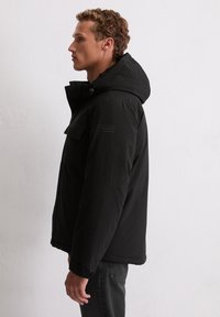 Zwarte donsjas met capuchon, voorzien van een voorklepzak en een logo badge, gemaakt van een glad, mat stof. Zijaanzicht weergegeven.