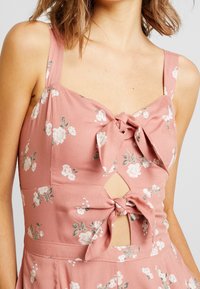 Frau in einem ärmellosen rosa Blumenkleid mit zwei vorderen Schleifen und Ausschnitten an Brust und Taille.