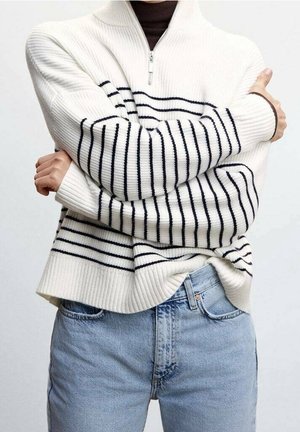 Person iført en hvid ribstrikket sweater med sorte vandrette striber og lyseblå jeans, med armene over kors foran brystet.