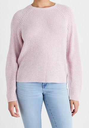 Personne portant un pull en maille texturée rose clair et un jean bleu clair, vu des épaules jusqu’aux hanches sur un fond uni.