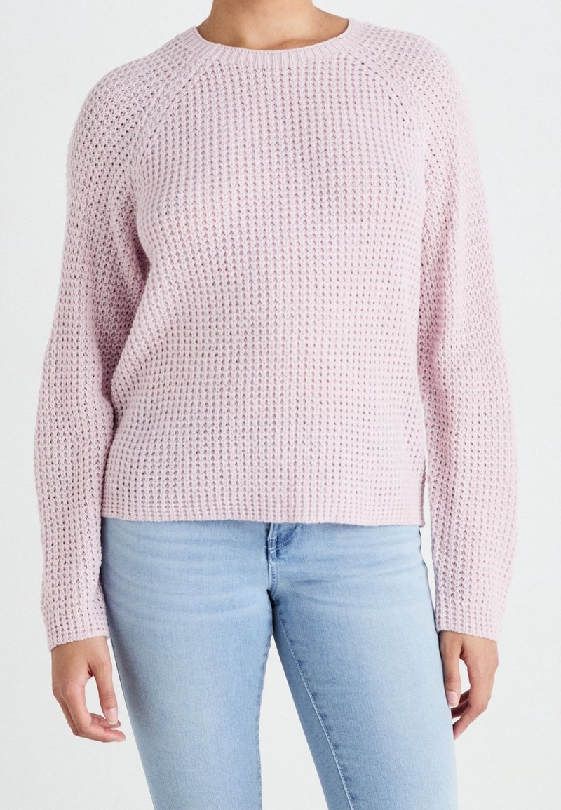 Personne portant un pull en maille texturée rose clair et un jean bleu clair, vu des épaules jusqu’aux hanches sur un fond uni.