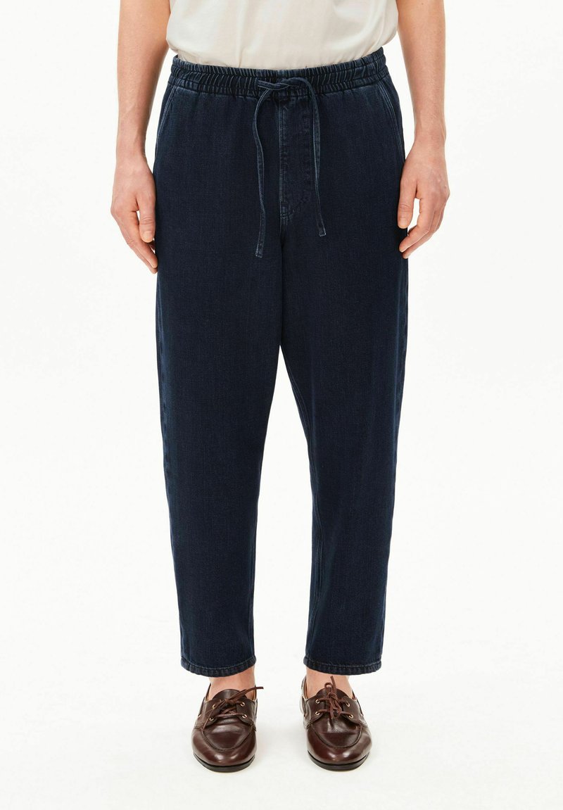 Personne portant un pantalon bleu foncé à cordon, une chemise blanche et des chaussures en cuir marron, debout devant un fond blanc uni.