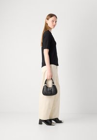 Borsa a mano in pelle nera con una forma strutturata, dettagli in metallo argentato, tenuta da una persona che indossa un top nero e pantaloni larghi color beige chiaro.
