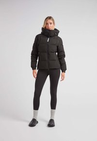 Zwarte pufferjack met een hoge kraag, een rits aan de voorkant en grote gestikte secties. Gestyled met zwarte leggins en grijze sokken.