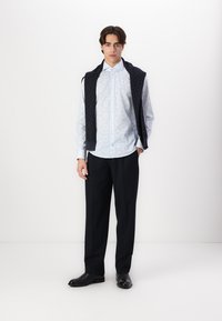 Lichtblauwe blouse met patroon en lange mouwen, met een button-down kraag, gecombineerd met donkere broek en een donkere gebreide vest over de schouders.