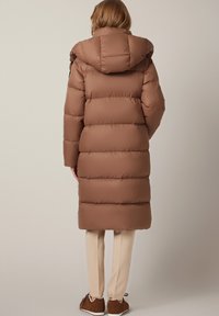 Brązowy puchowy płaszcz z kapturem, z poziomym przeszyciem, luźnym krojem i średniej długości. Połączony z jasnobeżowymi spodniami.