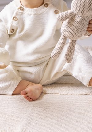 Tout-petit assis sur un tapis clair portant une tenue en tricot blanche, tenant un jouet en peluche beige au crochet.