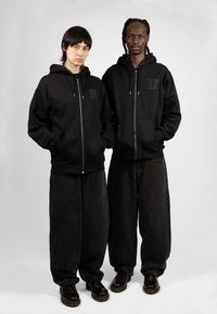 Sudaderas con cremallera negra con bolsillos frontales, que presentan un logo sutil. Combinadas con jeans oscuros de corte holgado y zapatos negros. Fondo neutro.