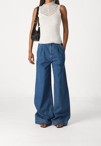 Bílé top bez rukávů s krajkovým lemem, zkombinované s volnými modrými džíny širokého střihu. Outfit doplňuje černá kabelka a sandálky na podpatku.